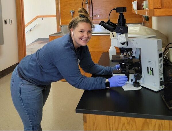 Anna stands at the microscope, wearing blue gloves and smiling at the camera.